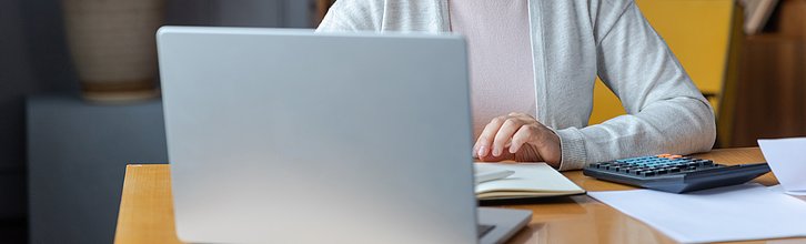Ein Schreibtisch auf dem ein Lapotop, Unterlagen und ein Taschenrechner liegen. Dahinter sitz eine Person.