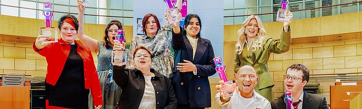 Gruppenbild der acht Preisträger*innen. Stehen jubelnd im Plenarsaal und halten die Auszeichnungen in die Höhe.