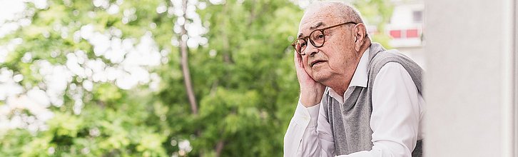 Älterer Person auf einem Balkon, die in die Ferne schaut. Im Hintergrund ein Baum.