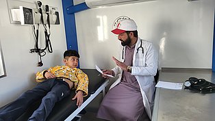 Innenaufnahme des LKW: Ein Erwachsener in Arztkittel spricht mit einem Jungen, der auf der Behandlungsliege liegt.