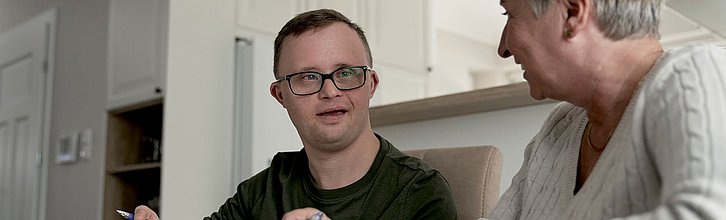 Eine Person mit Down-Syndrom sitzt mit Stift in der Hand neben einer anderen Person, die ihr freundliche zugewandt sitzt.