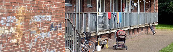 Eine heruntergekommene Hausfassade, davor steht eine Schubkarre und ein Kinderwagen. Über einem Geländer hängen Handtücher.