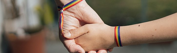 Zwei Hände halten sich. Beide tragen Armband in Regenbogenfarben.