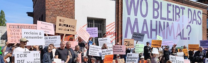 Menschen mit Demo-Schildern vor einem Banner mit der Aufschrift "Wo bleibt das Mahnmal".