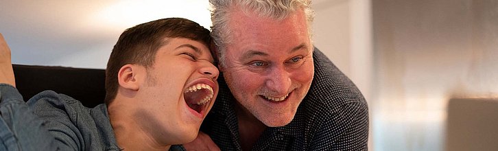 Ein lachender Jugendlicher mit Behinderung wird von einer lachenden Erwachsenen Person umarmt.