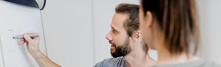 Eine Person steht mit Stift in der Hand am Flipchart und zeigt einer anderen Person etwas darauf.