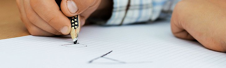 Kinderhand, die einen Buntstift hält und auf Papier ein großes und ein kleines "a" schreibt.