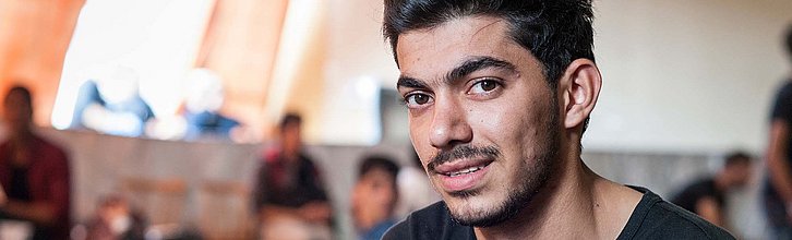 Eine Person mit schwarzen Haaren und Bart schaut in die Kamera. Im Hintergrund sind weitere Personen und Betten zu sehen.t