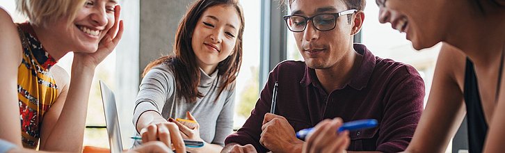 Vier Personen sitzen mit Schreibutensilien um einen Tisch herum und tauschen sich aus.