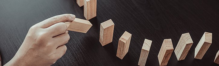 In eine Dominostein-Reihe aus Holzklötzchen wird ein fehlendes Klötzchen hinzugestellt.