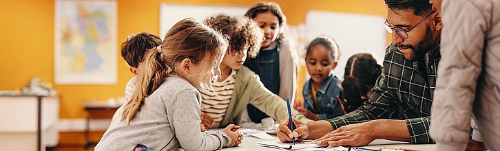 Ein Erwachsener, mehrere Kinder, alle schauen auf ein Blatt, auf das Erwachsener etwas schreibt