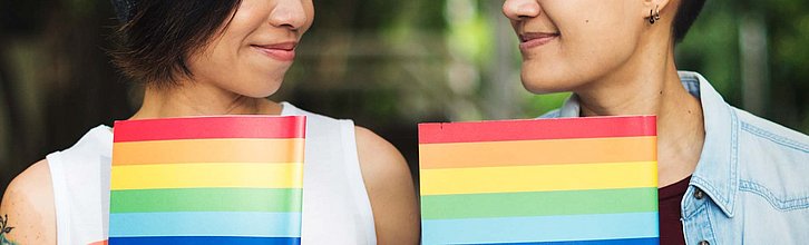 Zwei Personen, deren Gesichter nur zu Hälfte zu sehen sind und die sich anlächeln, halten Regenbogenfahnen