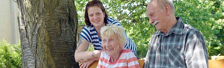 Zwei ältere Personen sitzen draußen auf einer Bank, eine jüngere Frau steht dahinter