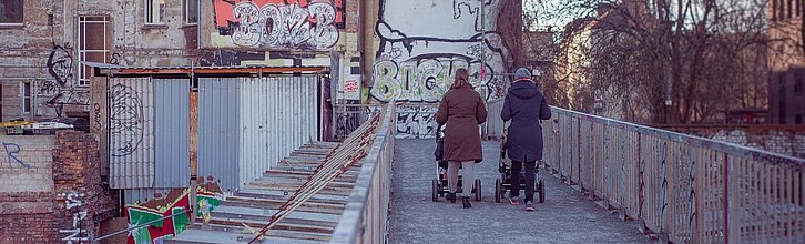 Zwei Personen mit Kinderwagen laufen durch ein heruntergekommens Wohnviertel mit Graffitti an den Wänden.