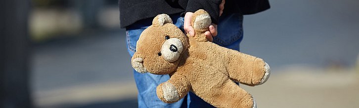 Ein Kind hält einen Teddy-Bären mit einer Hand hinter dem Rücken.