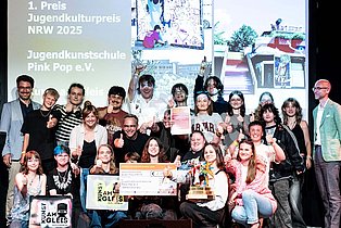 Die jugendlichen Gewinner*innen von Pink Pop halten den Preis, einen Scheck, und Logo-Plakate in die Kamera und jubeln auf der Bühne.