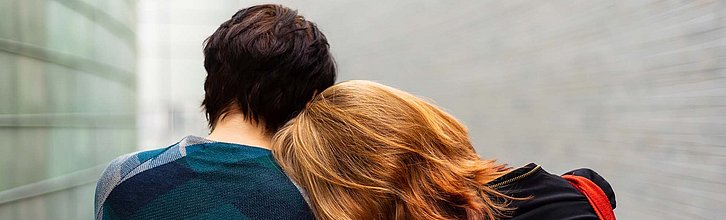 Rückansicht von zwei Personen. Die eine Person legt den Kopf auf die Schulter der anderen.