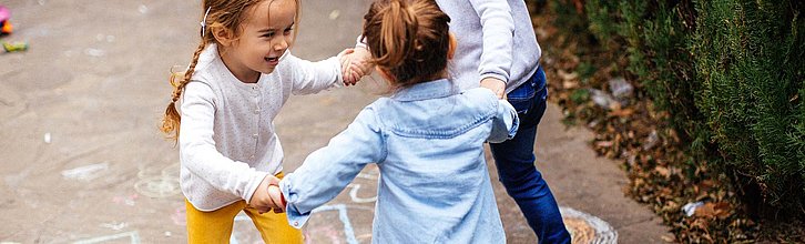 Drei Kinder, die sich an den Händen haltend im Kreis laufen und dabei glücklich aussehen.