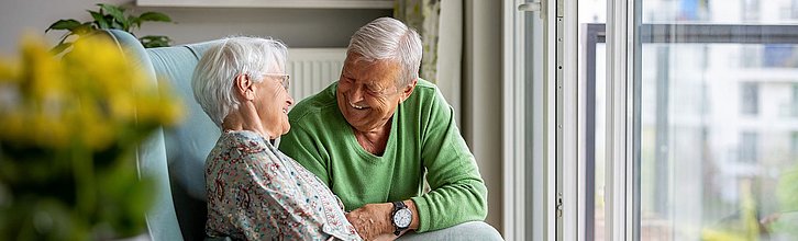 Eine ältere Person hockt neben einer anderen älteren Person, die in einem Sessel sitzt. Beide sind einander zugewandt und lächeln.