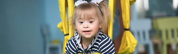Ein Kind mit Down-Syndrom hängt in einer Therapieschaukel und guckt in die Kamera.