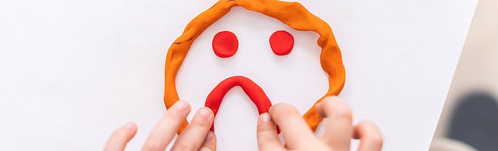 Kinderhände, die aus Knete ein trauriges Gesicht auf einem Tisch formen.