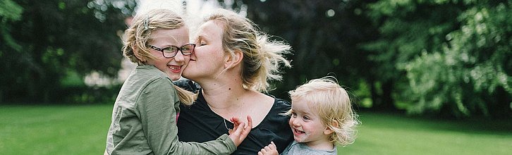 Eine Mutter auf einer Wiese mit zwei Kindern auf dem Arm. Alle drei lachen und sehen glücklich aus.