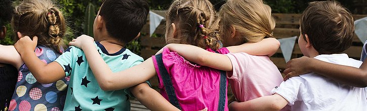 Mehrere Kinder von hinten fotografiert, die ihre Arme umeinander legen