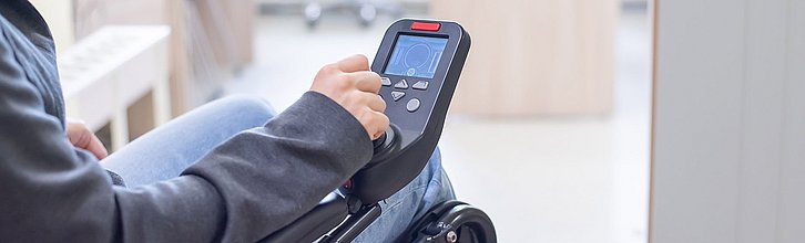 Hand einer im Rollstuhl sitzenden Person umfasst einen Steuerknüppel.