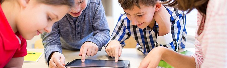 Drei Kinder sitzen über Tablets gebeugt. Eine Erwachsene Person sitzt daneben.