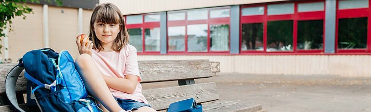 Ein Kind sitzt neben seinem Schulranzen auf einer Bank mit einem Apfel in der Hand. Im Hintergrund ein Schulgebäude.