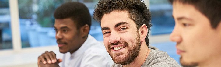 Drei junge Erwachsene sitzen in einem Seminarraum. Eine Person lächelt glücklich in die Kamera.