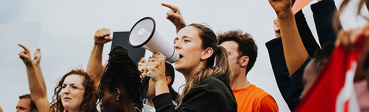 Eine Gruppe junger Menschen demonstriert. Eine Frau in der ersten Reihe ruft etwas in ein Megafon.