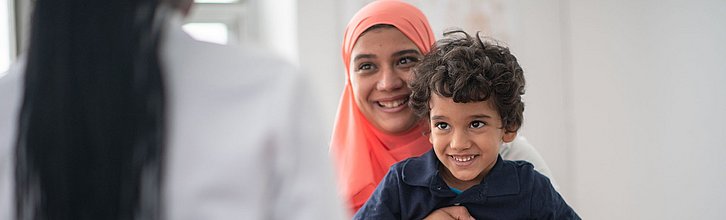 Eine Frau, die Kopftuch trägt und ein Kind auf dem Schoß hat, sitzt einer anderen Person gegenüber und lächelt.