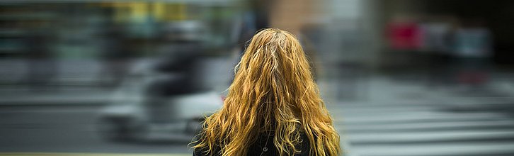 Person mit langen Haaren steht an einem Zebrastreifen. Der Hintergrund ist verschwommen.