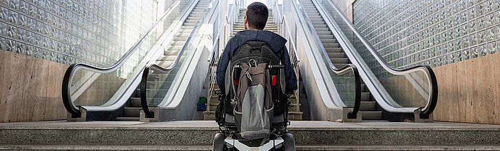 Person im Rollstuhl vor einem U-Bahn-Aufgang mit Treppen und Rolltreppen.