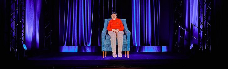 Hologramm auf einer Bühne, das eine ältere Person zeigt, die auf einem Sessel sitzt.