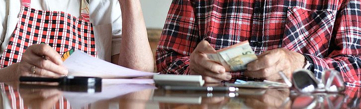 Oberkörper von zwei Personen, die an einem Tisch sitzen. Eine Person hält kleine Geldscheine in der Hand, die andere einen Zettel.