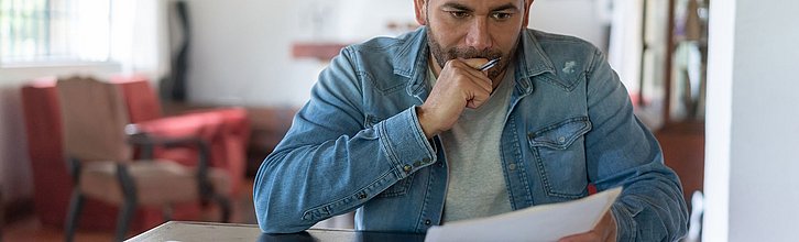 Eine Person sitzt mit ratlosem Blick vor einem Tisch voller Dokumente.