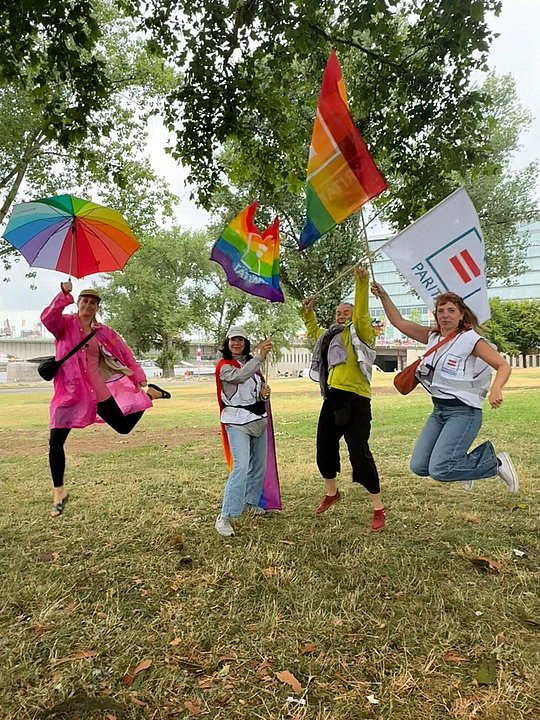 Vier Personen springen in die Luft. Sie halten Regenbogenfahnen, Schirme und Fahnen des Paritätischen.