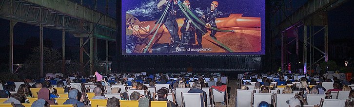 Eine Filmvorführung. Viele Menschen sitzen auf Stühlen vor einer Leinwand. Auf der Leinwand ist ein Filmausschnitt zu sehen.