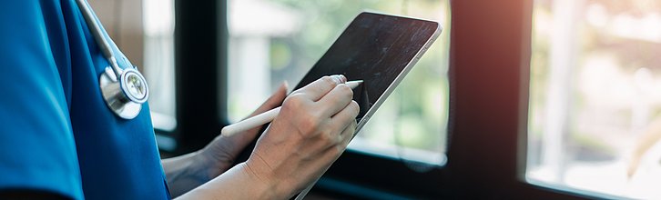 Hand, die etwas auf ein Tablet schreibt. Zu sehen ist auch ein Kasock und ein Stethoskop