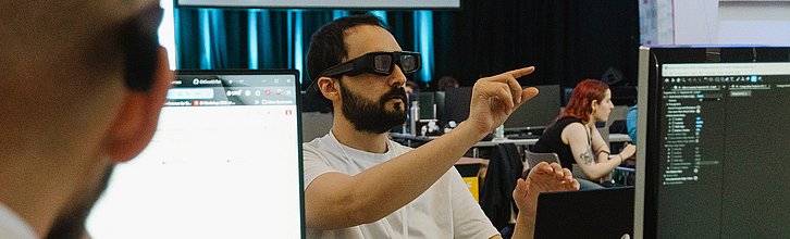 Eine Person hat eine VR-Brille auf und gestikuliert. Zwei weitere Personen sitzen vor Computerbildschirmen.