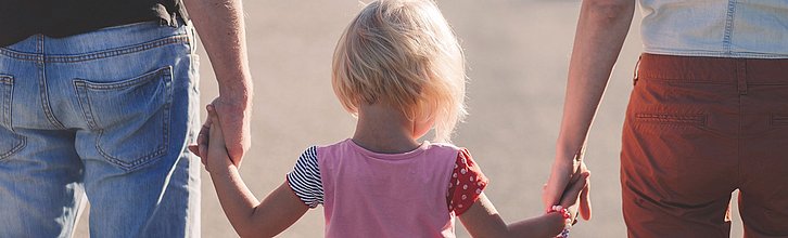 Rückansicht von zwei Erwachsenen, die in der Mitte ein Kind an den Händen halten.