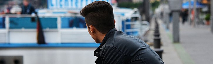 Person mit schwarzen Haaren sitzt auf einem Pöller in einer Fußgängerzone und schaut in die Ferne.
