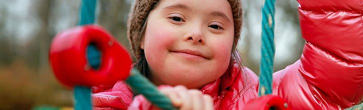 Ein Mädchen mit Trisomie 21 trägt eine rote Jacke und spielt auf einem Klettergerüst und lächelt in die Kamera.