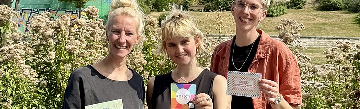 Drei Personen vor grünen Büschen. Person in der Mitte hält den Respect!-Aufkleber in die Kamera