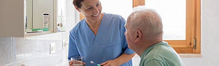 Pflegefachkraft steht neben einer älteren Person am Waschbecken mit Zahnbürste in der Hand und lächelt ihr zugewandt zu.