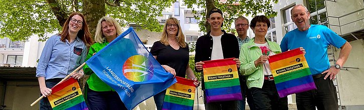 Gruppenbild von sieben Personen, die regenbogenfarbene Sportbeutel und eine Fahne mit Regenbogenlogo halten.