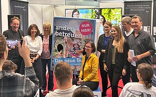 Sieben Personen, die neben einem Roll-Up, das das Buch-Cover zeigt, stehen. Einer Person sitz dabei.