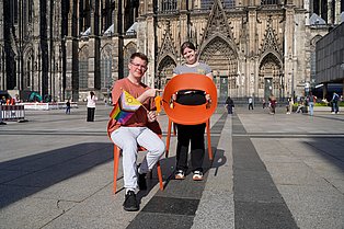 Zwei junge Erwachsene vor dem Kölner Dom. Eine Person sitz auf einem Stuhl, die andere steht mit einem Stuhl in den Händen daneben.